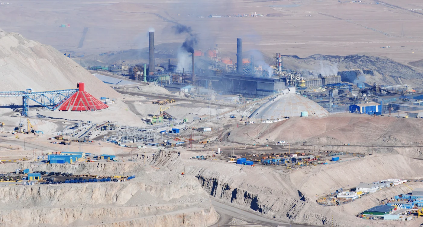 Fotografía referencial: chimeneas y edificios de una fundición de cobre entre correas transportadoras y acopios en el desierto de Atacama.