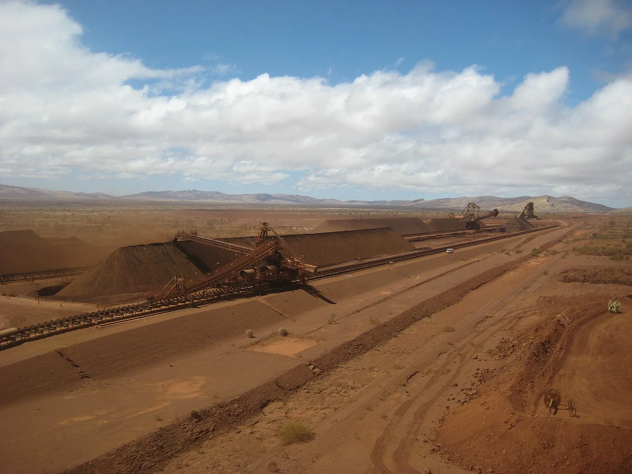 Fotografía referencial: apilador y correas transportadoras junto a largos acopios de mineral en un paisaje minero árido.