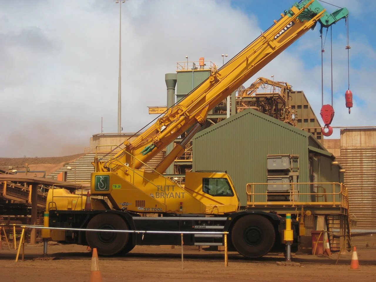 Fotografía referencial: grúa móvil amarilla junto a estructuras metálicas y equipos de una planta de procesamiento de mineral sobre terreno seco.