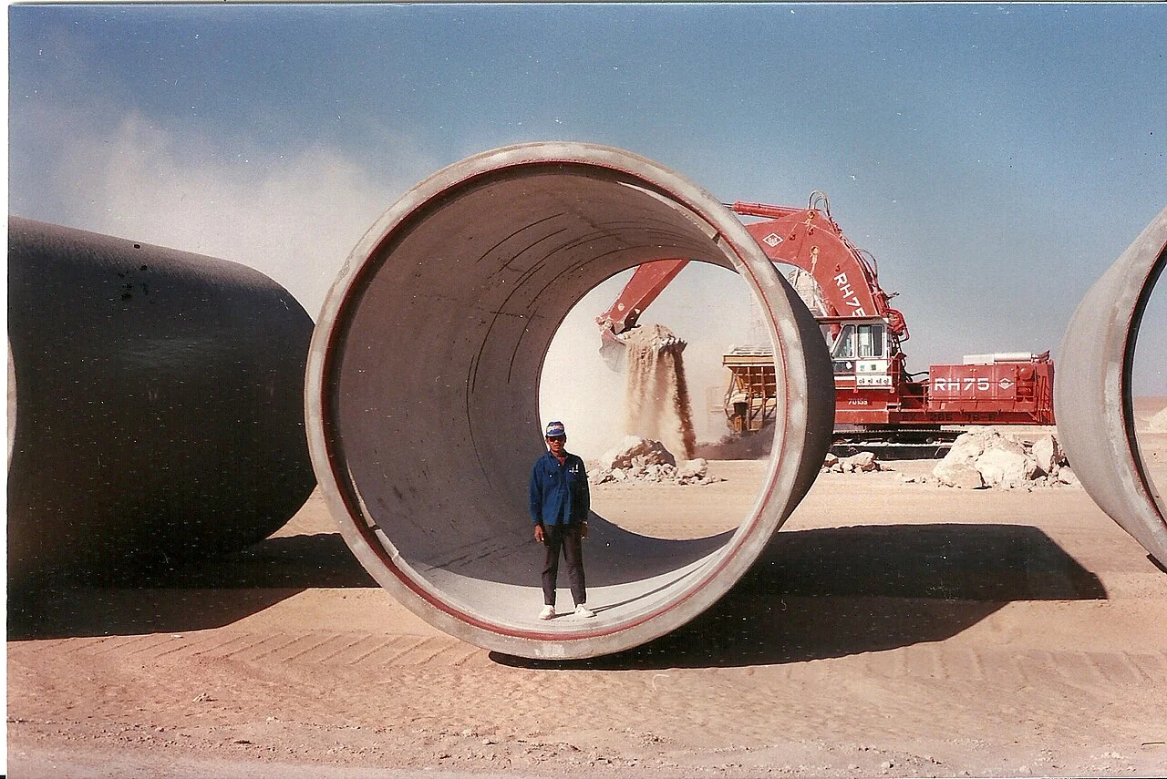 Fotografía referencial: trabajador de pie dentro de una tubería de gran diámetro, con excavadora y movimiento de tierras en un desierto.